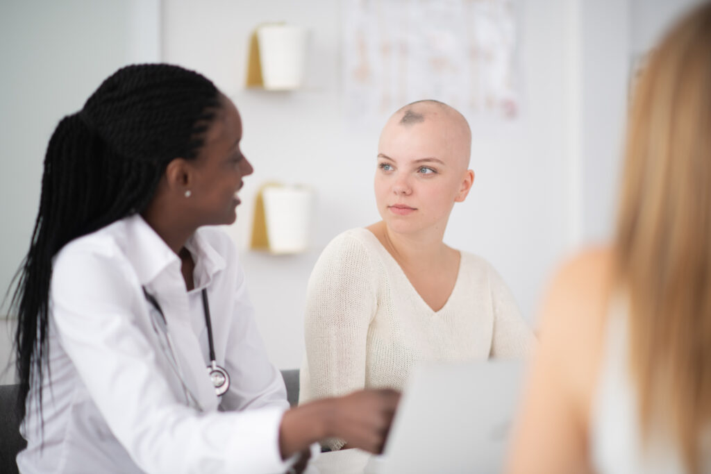 A woman is indoors in a hospital room. Her head is shaved due to chemotherapy. She is listening to her doctor give a prognosis.